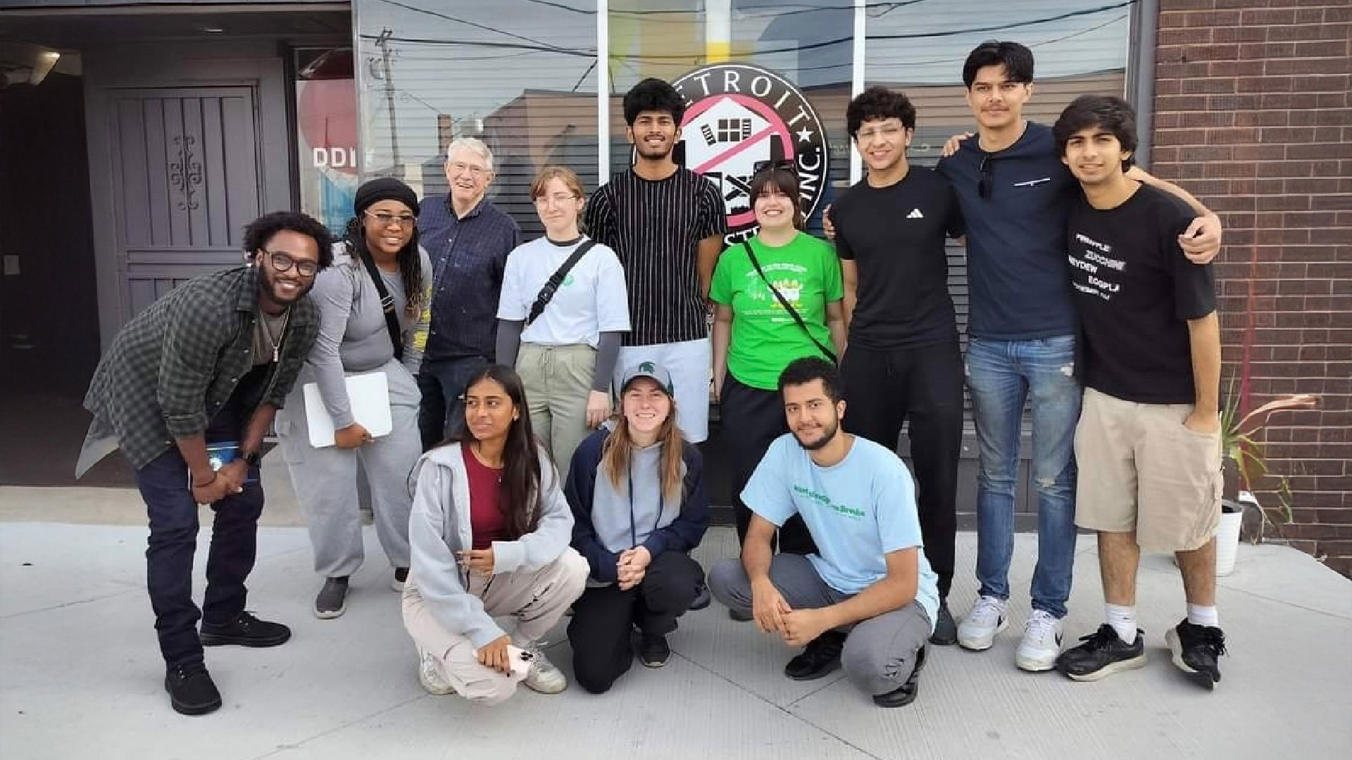 A group of people poses together outside a building with a large circular sign that reads “Detroit” and includes a graphic of a house. The group is standing and kneeling on a sidewalk in front of a brick wall and glass doors.