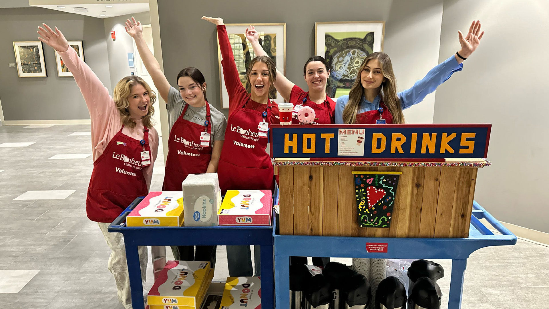 A group of volunteers wearing red aprons stands behind a wooden cart labeled “HOT DRINKS.” The cart has a festive design and is stocked with beverage supplies and boxes. The volunteers have their arms raised in a cheerful pose in a brightly lit hallway with framed artwork on the walls.
