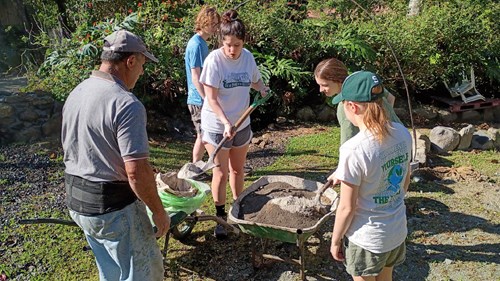 Several individuals are working outdoors in a lush green area with plants and rocks. One person is shoveling material into a wheelbarrow while others stand nearby observing or assisting. The setting appears to be a garden or restoration site.