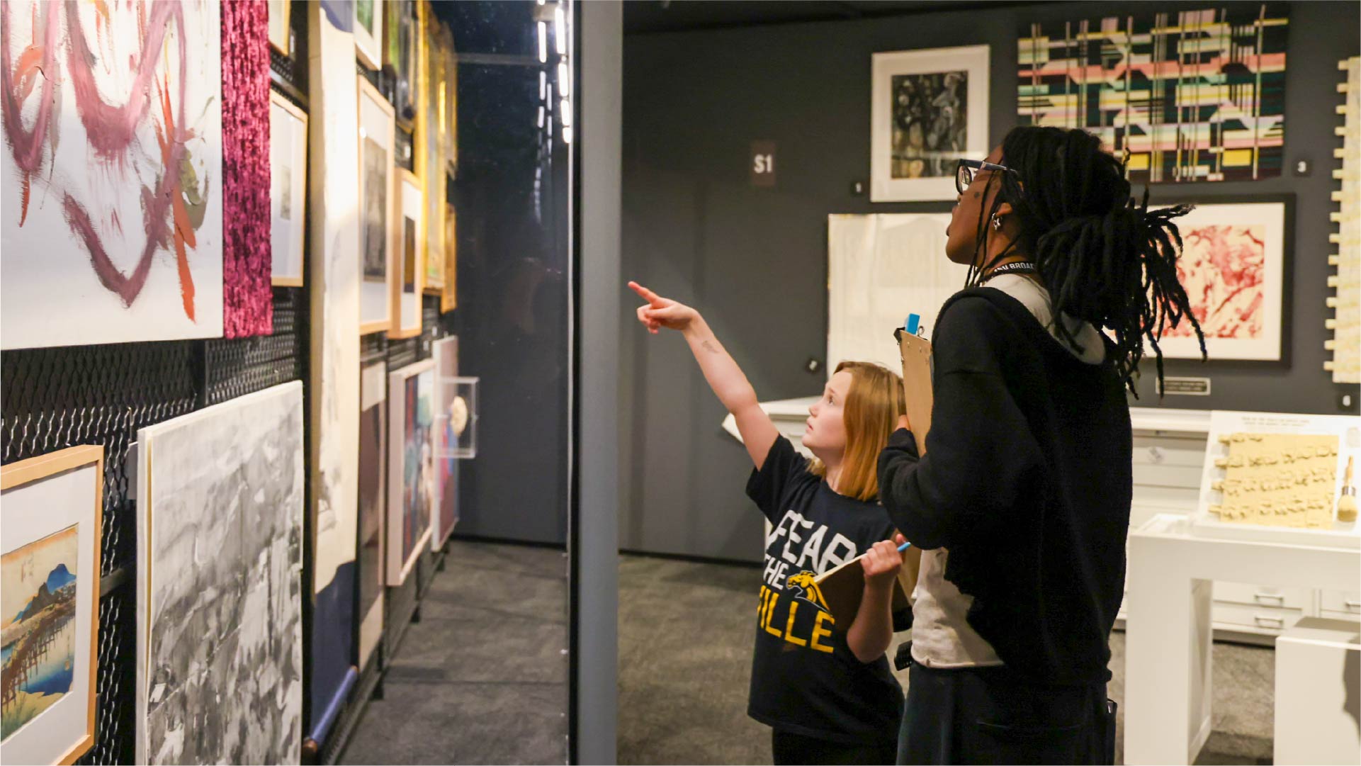 A classroom filled with young students seated at desks, many raising their hands. Two presenters stand at the front near a whiteboard and a large screen displaying a slide with text and images. The classroom walls are decorated with colorful posters and charts, and an American flag is visible near the board.