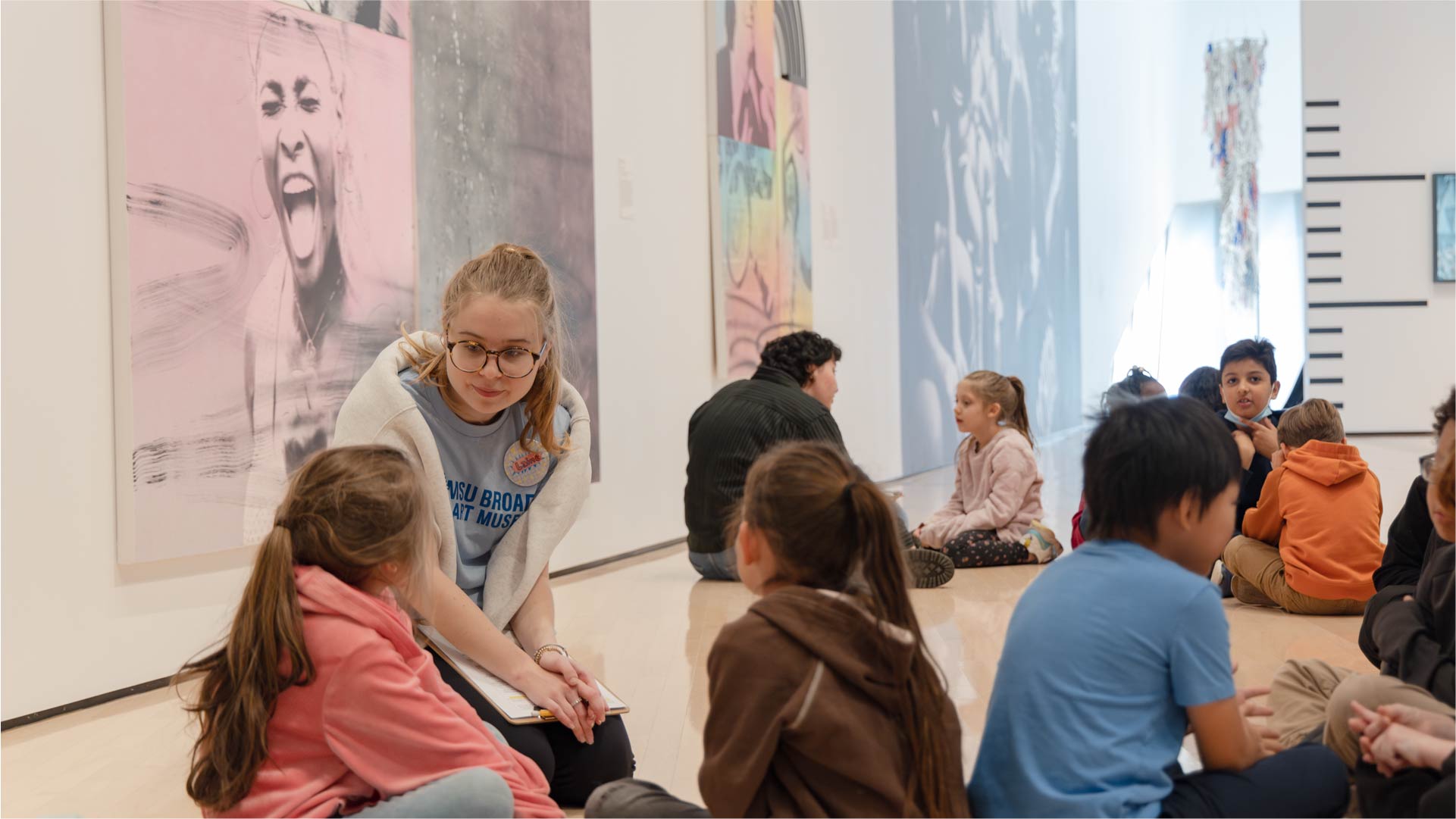 Several children sit on the floor in a museum gallery, engaged in conversation with a presenter. Large contemporary artworks, including a portrait and abstract pieces, are displayed on the walls behind them. Other small groups are visible in the background, also seated and interacting.