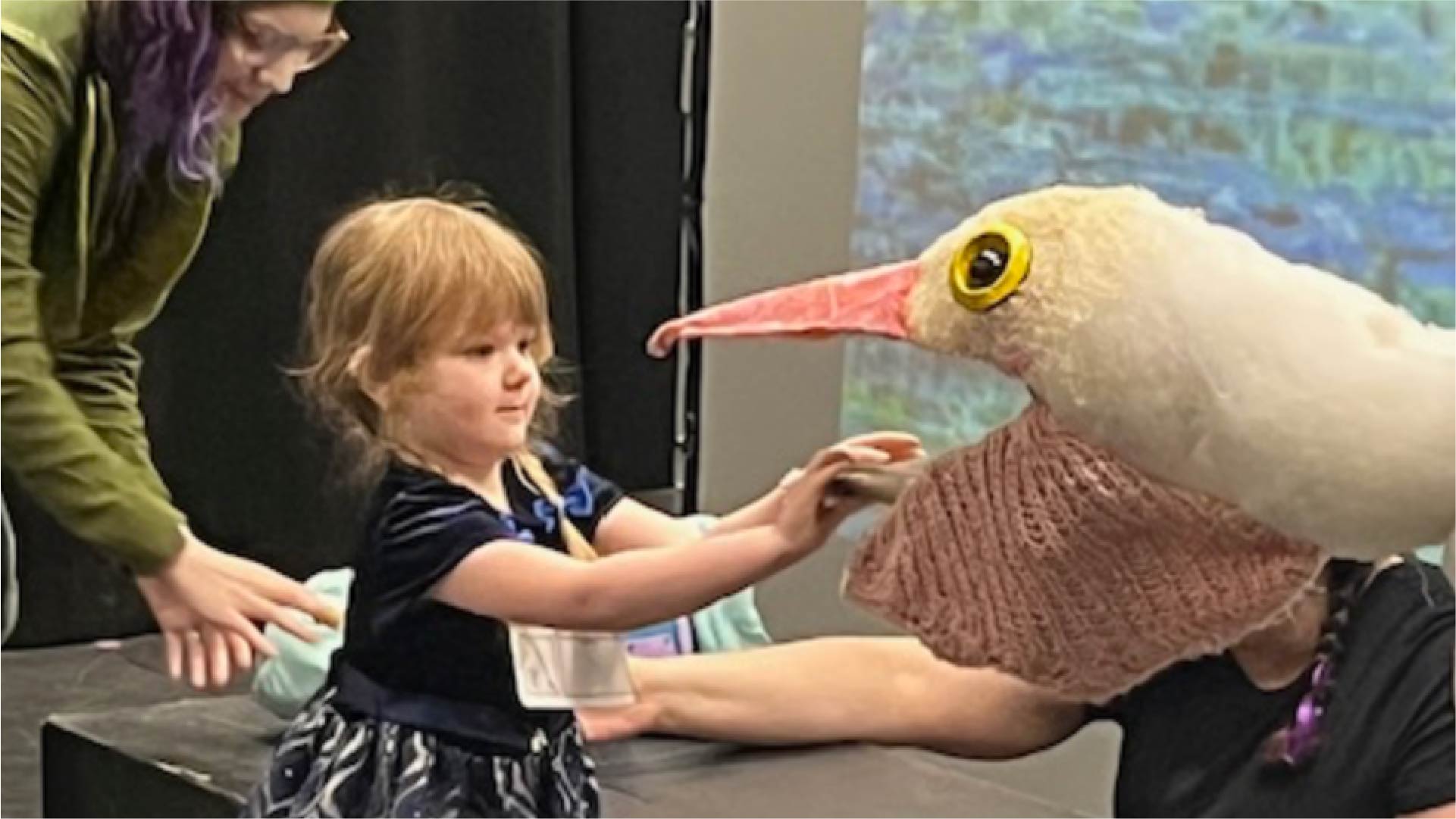 A child reaches out to touch a large bird puppet with a white head, yellow eye, and long pink beak. The puppet is held by a person wearing a black shirt, and another person stands nearby assisting. The background includes a projection screen displaying a colorful image.