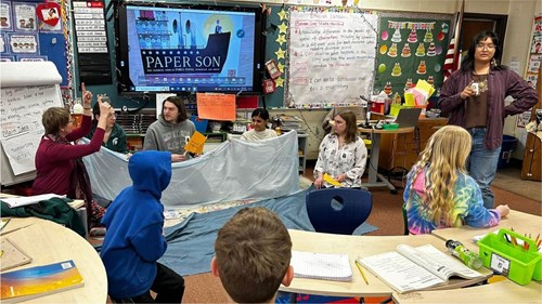 A classroom scene where students and two adults are engaged in an interactive activity. Several children sit at desks while others sit behind a light blue fabric sheet held up like a prop. A large monitor at the front displays the title “Paper Son” with an image of a ship. The walls are decorated with colorful charts, posters, and an American flag.