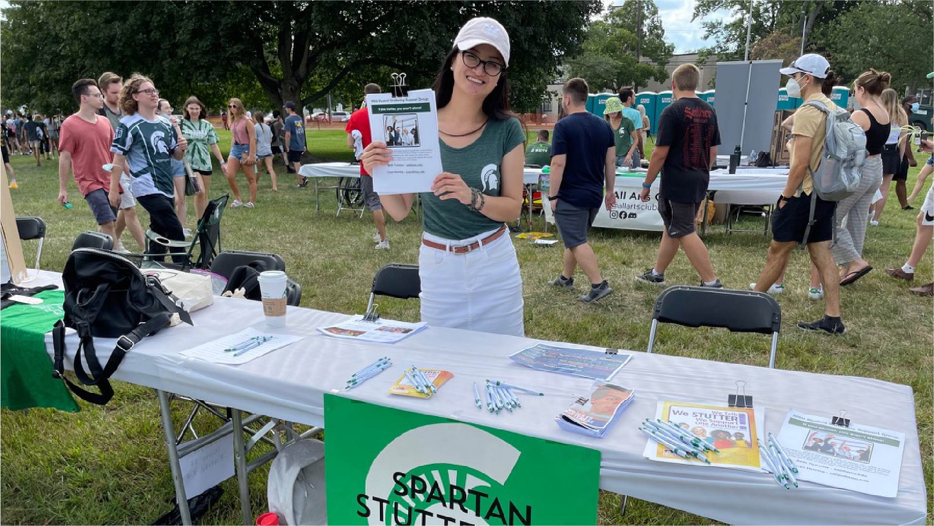 An individual stands behind a table at an outdoor event holding a flyer. The table is covered with informational materials, including brochures, pens, and a green sign that reads “Spartan Stuttering.” People are walking in the background, and the setting appears to be a campus fair or student organization event.