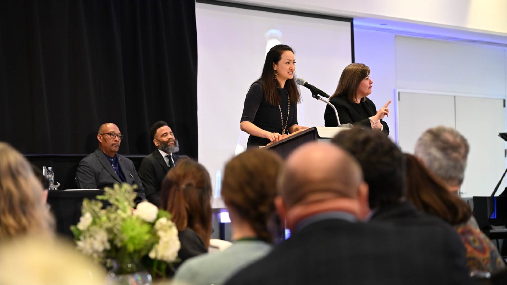 Two individuals stand at a podium speaking to an audience during an indoor awards ceremony. One person is at the microphone while the other gestures beside them. A black curtain and projection screen are in the background, and several people are seated facing the stage.