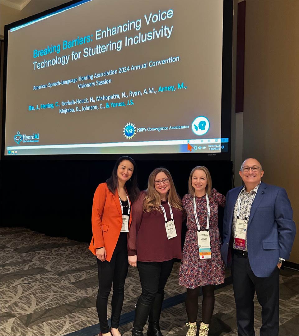 Four individuals stand in front of a large presentation screen at a conference. The screen displays the title “Breaking Barriers: Enhancing Voice Technology for Stuttering Inclusivity” along with the subtitle “American Speech-Language Hearing Association 2024 Annual Convention Visionary Session” and a list of authors. Logos for HeardAI and NSF’s Convergence Accelerator are visible at the bottom of the screen.