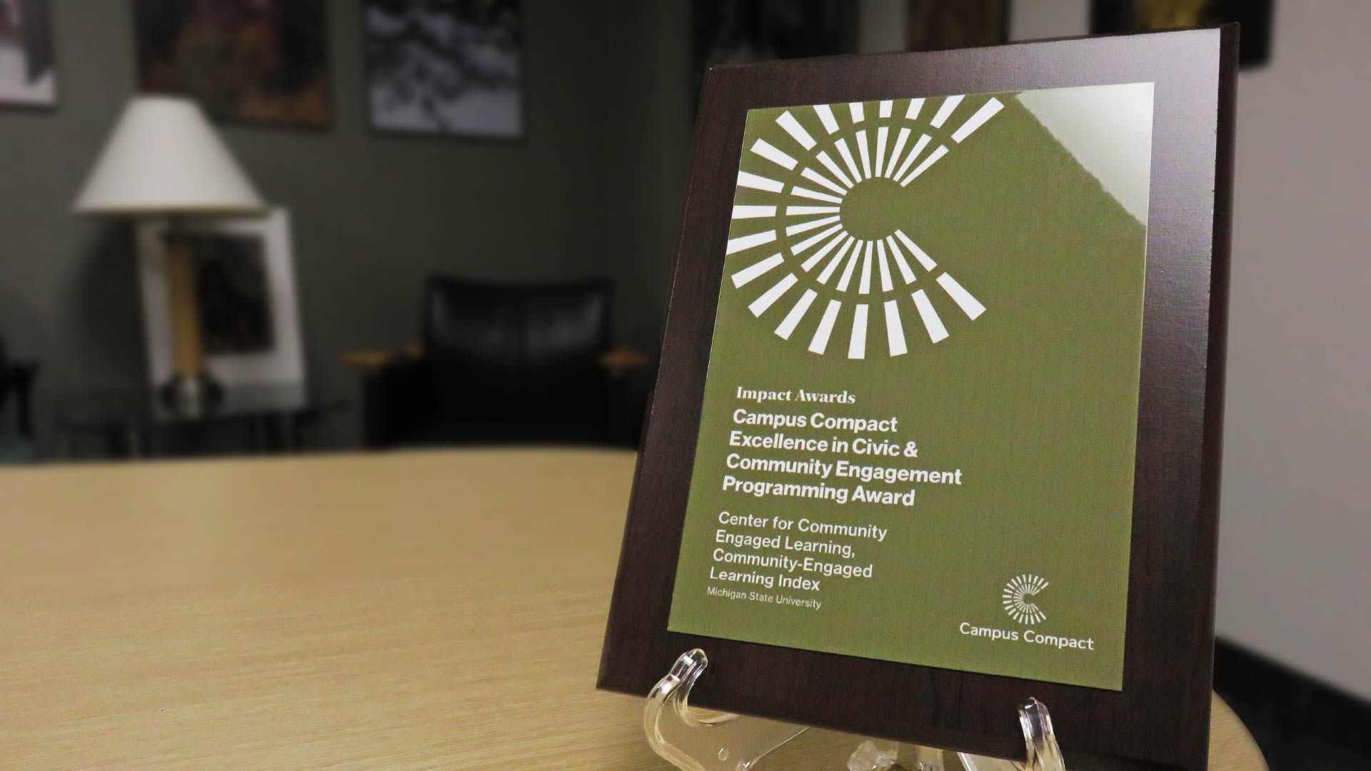 A close-up of a green and white award plaque on a clear stand. The plaque reads “Campus Compact Excellence in Civic & Community Engagement Programming Award” and includes the Michigan State University logo. The background shows a blurred office setting with framed artwork.