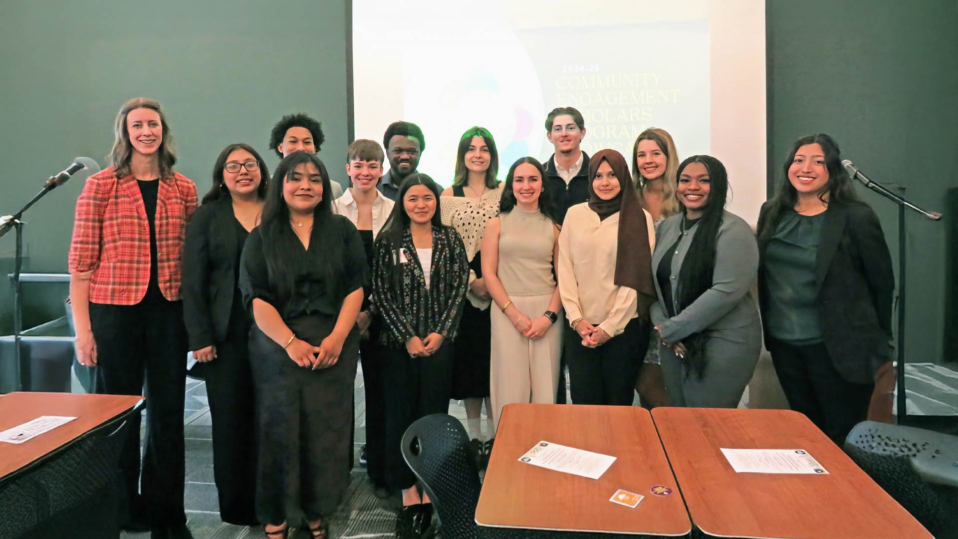 A group of people standing together in a conference room with two microphones on stands visible on either side. A presentation screen in the background displays text related to a community engagement scholars program. Tables with papers and chairs are in the foreground.