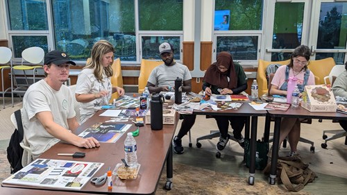 Several individuals seated around a table working on arts and crafts projects. The table is covered with magazines, scissors, glue, and water bottles. Large windows in the background show greenery outside.
