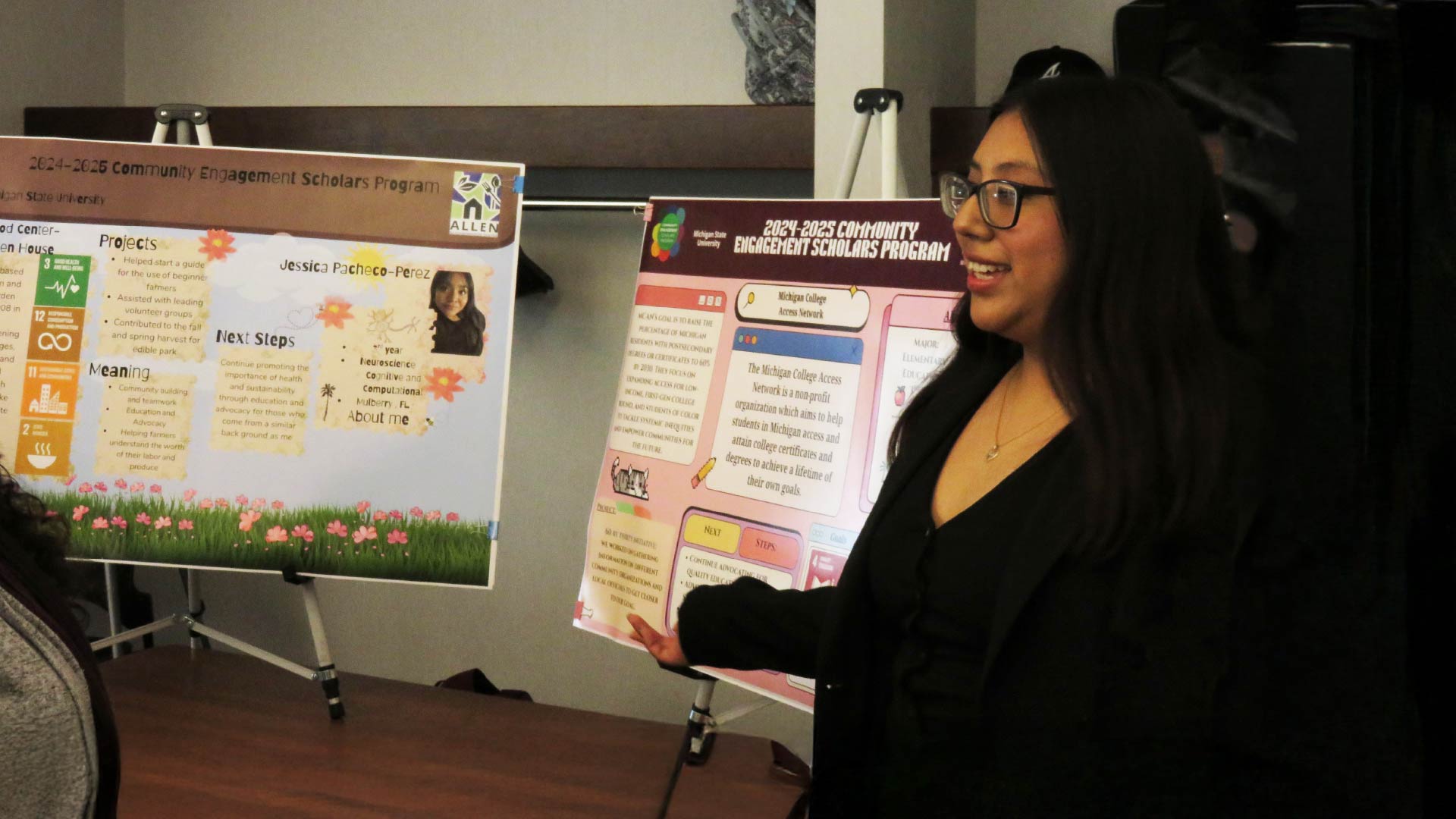 A person standing next to two colorful posters mounted on easels. The posters display information about community engagement projects, including graphics, text, and decorative elements.