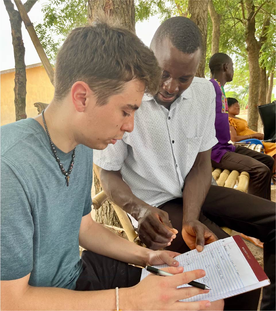 Two individuals seated on a wooden bench outdoors, reviewing a notebook with handwritten notes. One person holds a pen while the other points at the page. Additional people are visible in the background sitting under the shade of trees.