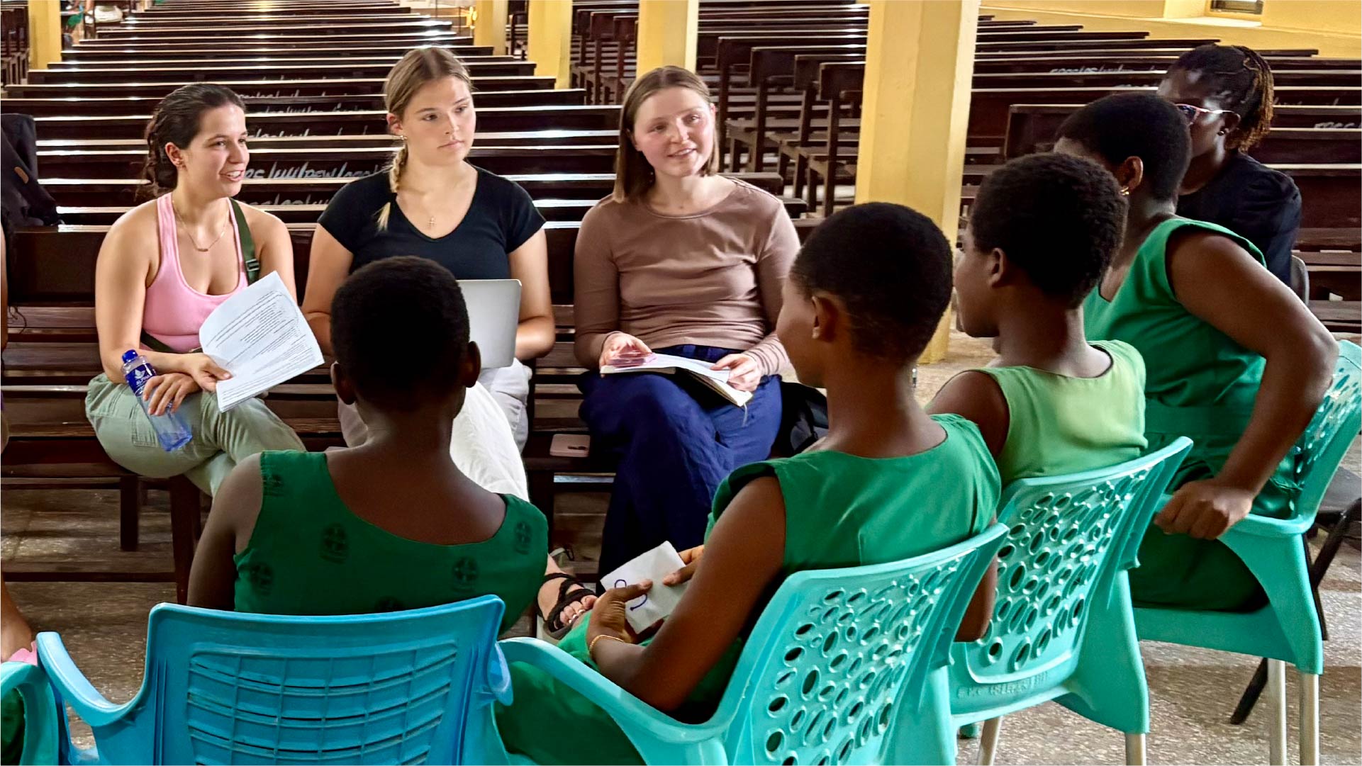 A small group of people seated in a circle inside a large hall with wooden pews. Three individuals hold papers and a laptop while engaging in conversation with several students wearing green uniforms seated on turquoise plastic chairs.