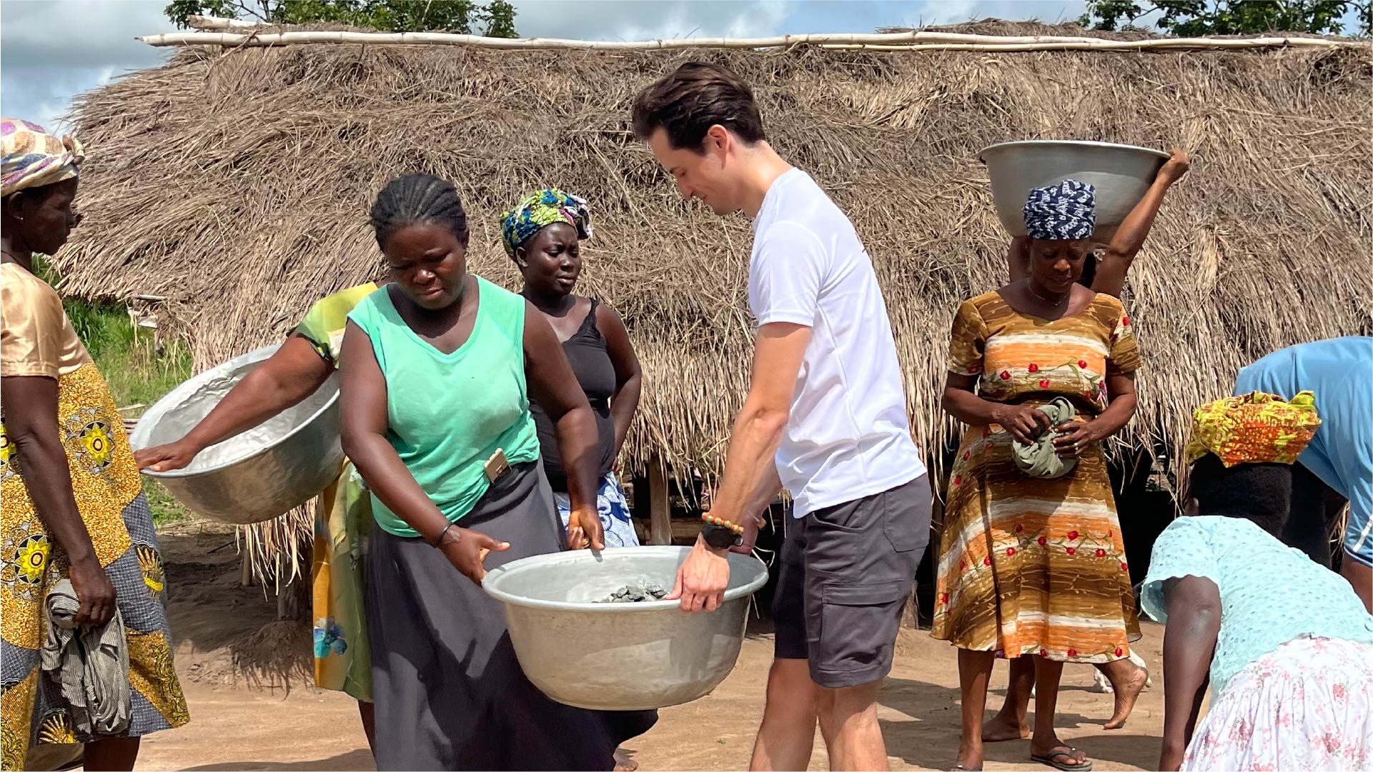 A group of people gathered outside near a thatched-roof structure. Several individuals carry large metal basins, and one person in a white shirt interacts with a community member holding a basin filled with items. Others stand nearby, some balancing basins on their heads.