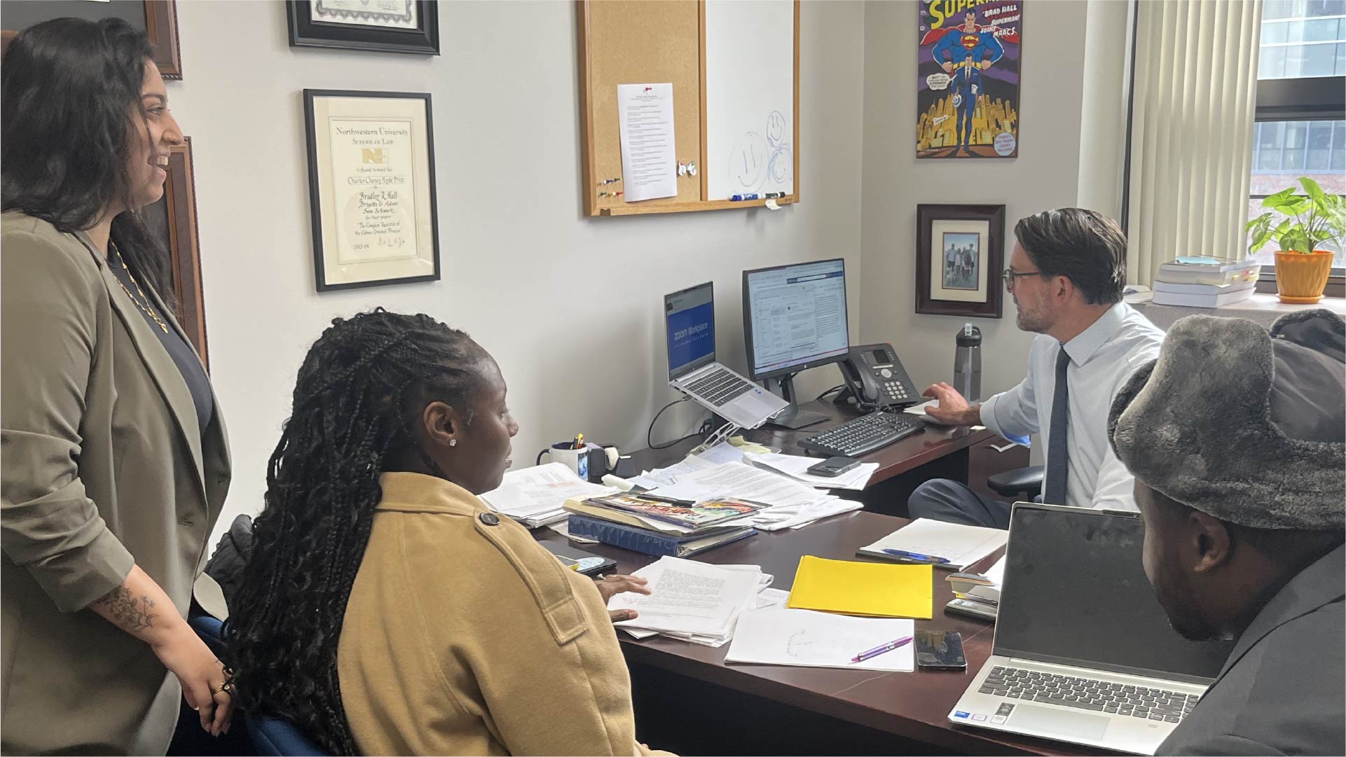 Four individuals are gathered in a small office around a desk covered with papers, folders, and a laptop. One person is seated at the desk pointing at a computer monitor displaying a document, while others stand or sit nearby observing. The office walls feature framed certificates, a corkboard, and a colorful poster.