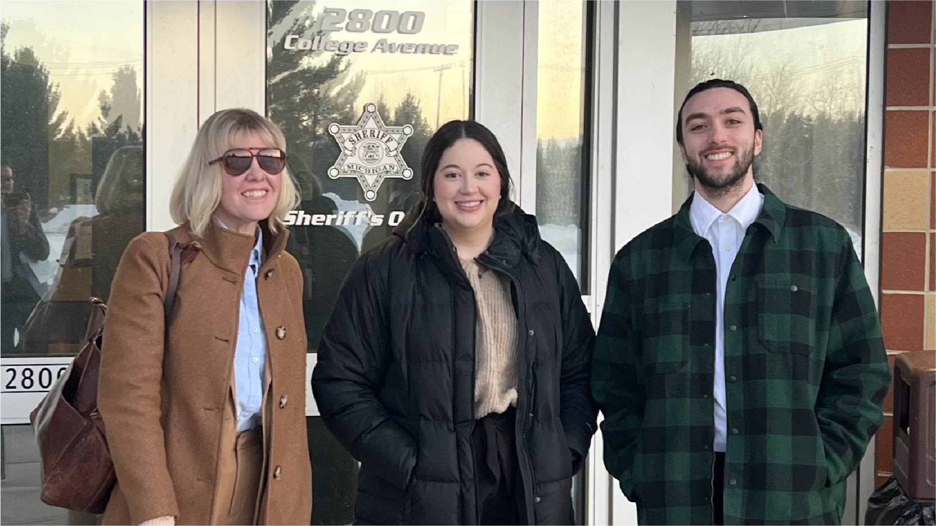 Three individuals stand in front of glass doors with signage that reads “Sheriff’s Office” and “2800 College Avenue.” The background includes reflections of trees and a clear sky.