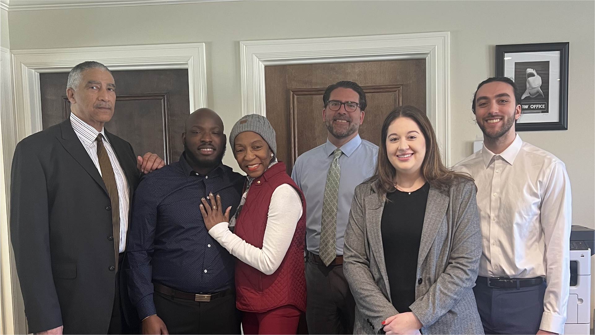 Six individuals stand together inside a room with light-colored walls and wooden doors. A framed picture hangs on the wall behind them, and a printer is visible in the corner.