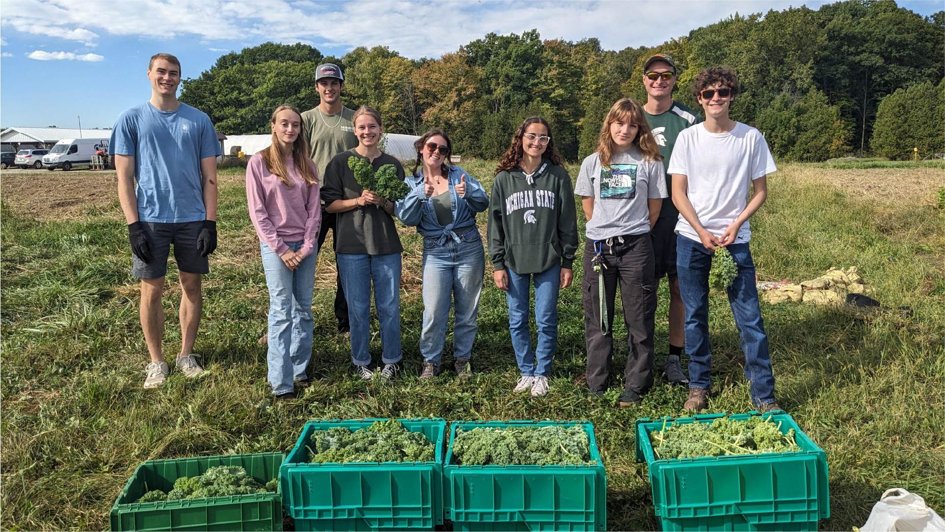 A group of eight individuals stands in a grassy field with trees in the background. In front of them are several green crates filled with leafy vegetables, likely kale. The setting appears to be a farm or garden area under a partly cloudy sky.