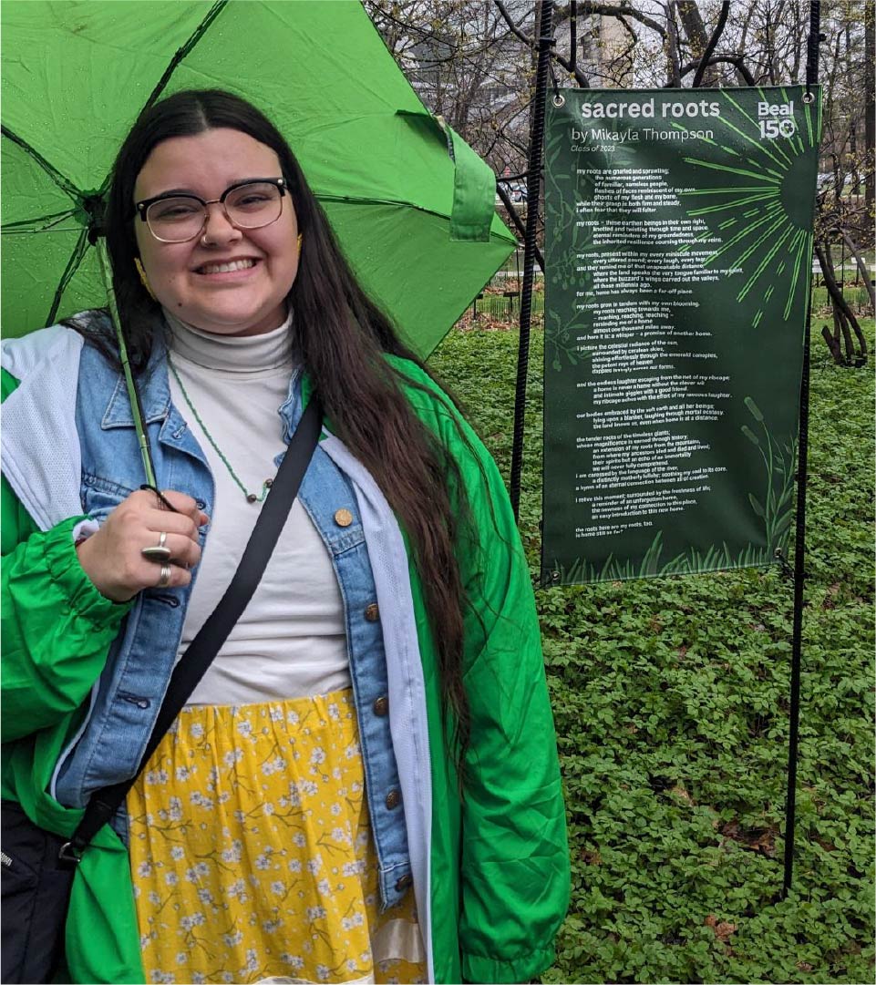 A person stands outdoors holding a green umbrella next to a tall sign titled “Sacred Roots by Mikayla Thompson.” The sign contains a poem printed in white text on a dark background with green decorative lines. Green plants and trees are visible in the background.