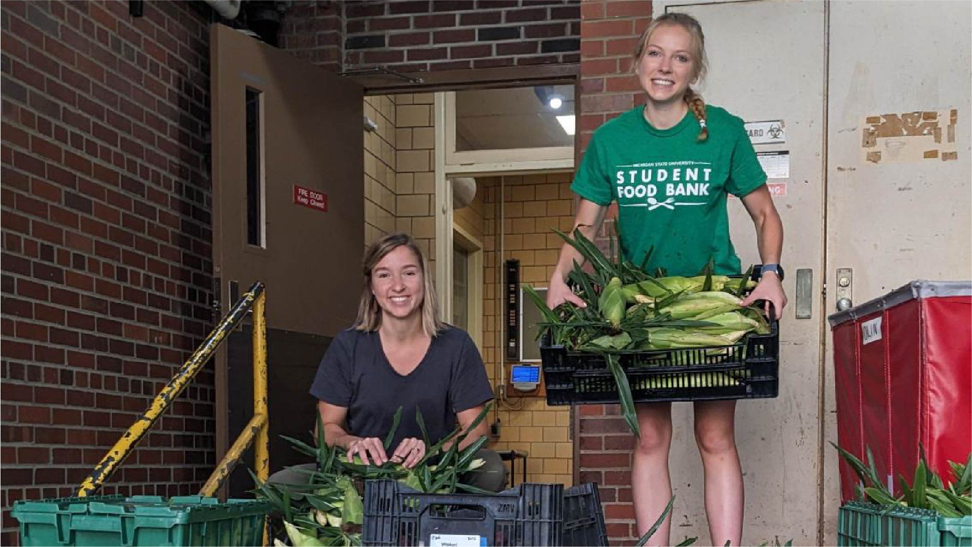Two individuals are handling fresh corn in black plastic crates outside a brick building. One person is seated, removing husks, while the other stands holding a full crate of corn. A green bin and a red container are nearby, and the person standing wears a green shirt with the words “Student Food Bank.”