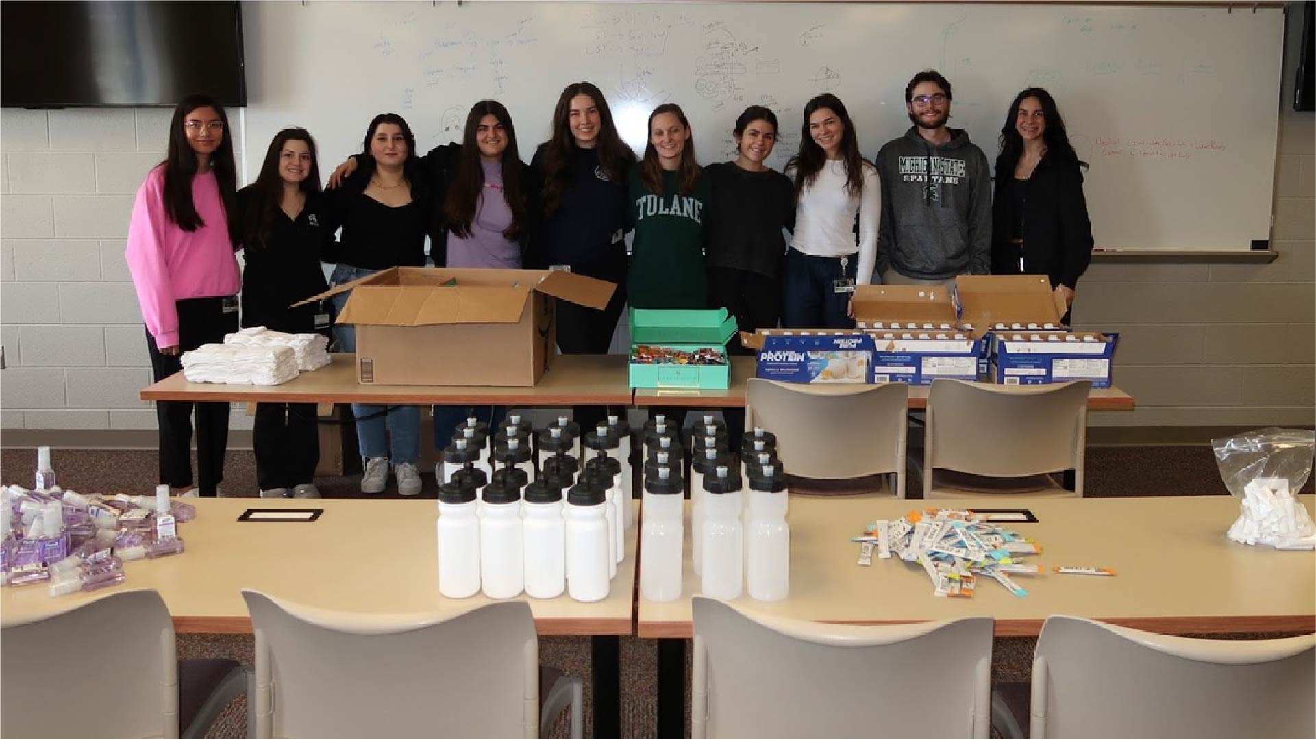 A group of people stands behind a long table filled with supplies for care kits. Items include water bottles, protein bars, toothbrushes, toothpaste, and hygiene products arranged in neat rows. Cardboard boxes and packaged goods are on the table, and a whiteboard with writing is visible in the background.