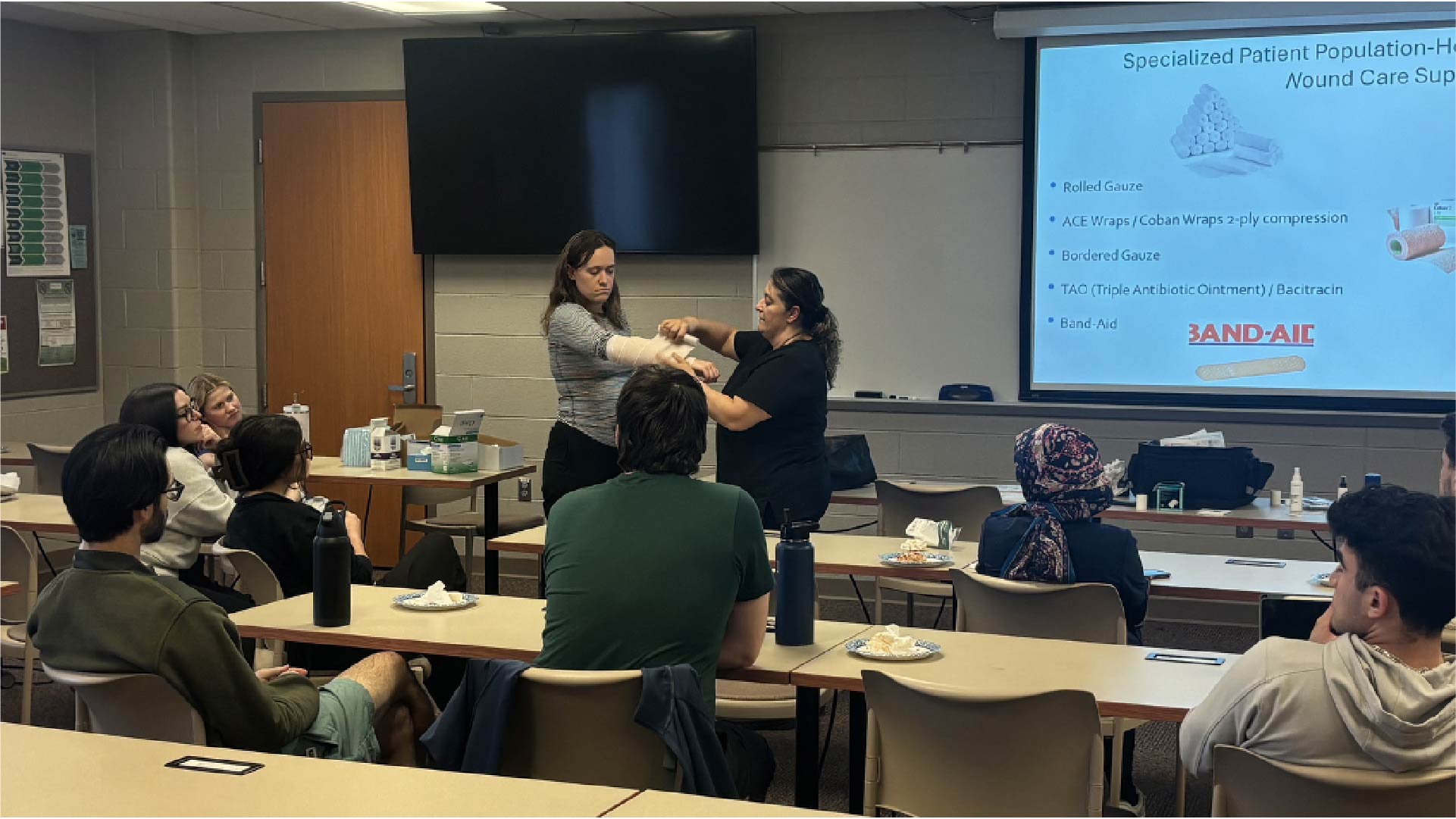 A classroom setting with several people seated at tables facing a presentation screen. Two individuals stand at the front demonstrating wound care techniques using bandages. The screen displays a slide titled “Specialized Patient Population – Wound Care Supplies” with a list of items including rolled gauze, ACE wraps, gauze pads, triple antibiotic ointment, and Band-Aid. Tables have water bottles and small supplies.