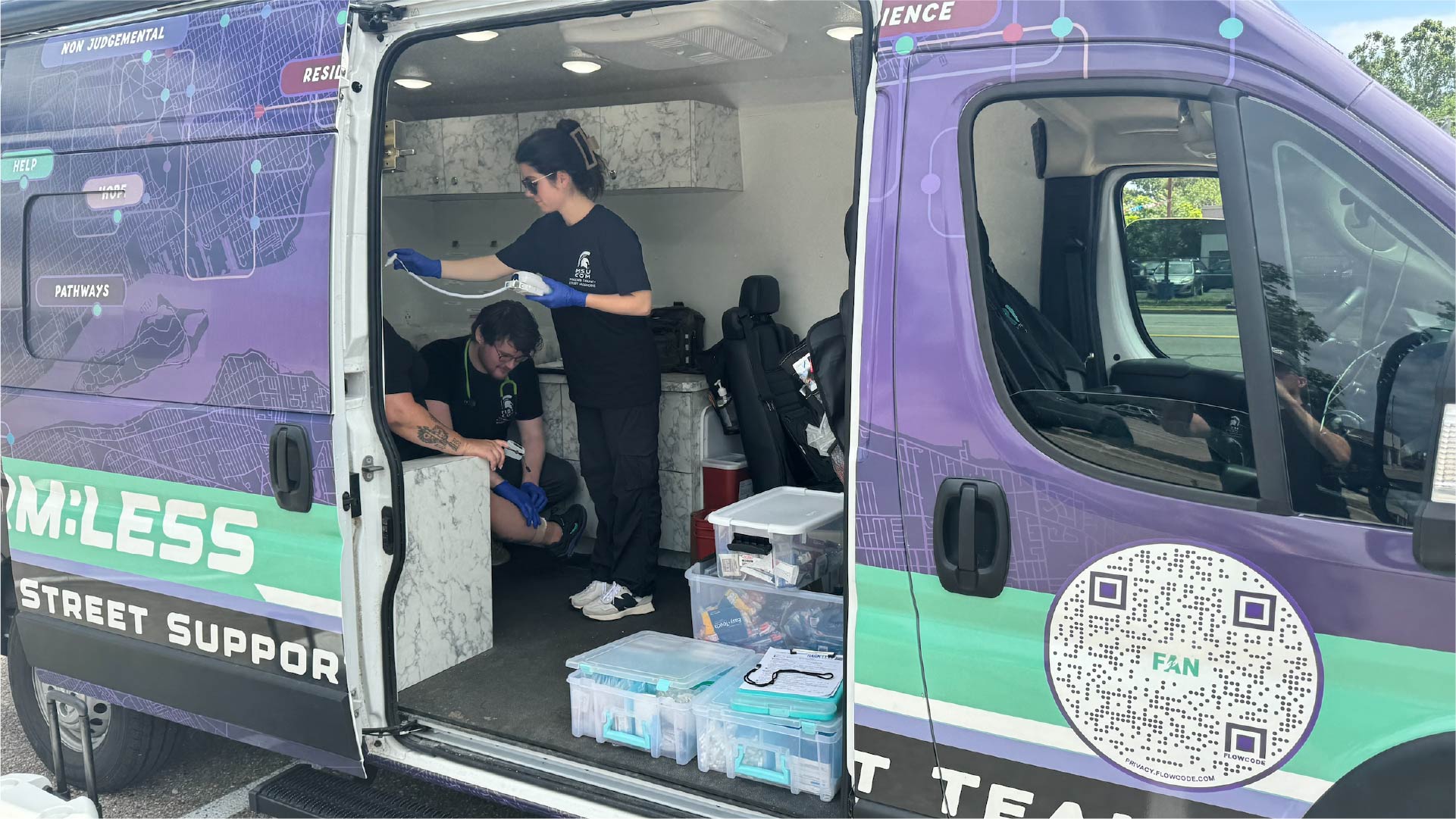 Inside a brightly colored mobile medical van, one person is seated while another applies care to the arm using gloves. The van’s interior includes storage compartments and clear plastic bins with medical supplies. The exterior of the van features a design with text and a QR code, along with words like “Team:LESS” and “Street Support.”