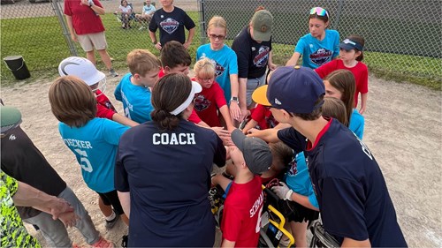 A group of youth baseball players wearing red and blue shirts gathers in a circle on a dirt field near a chain-link fence. All players have their hands stacked together in the center for a team cheer. One person wearing a navy shirt with the word “COACH” printed on the back is in the foreground. Baseball equipment and a few spectators are visible in the background.