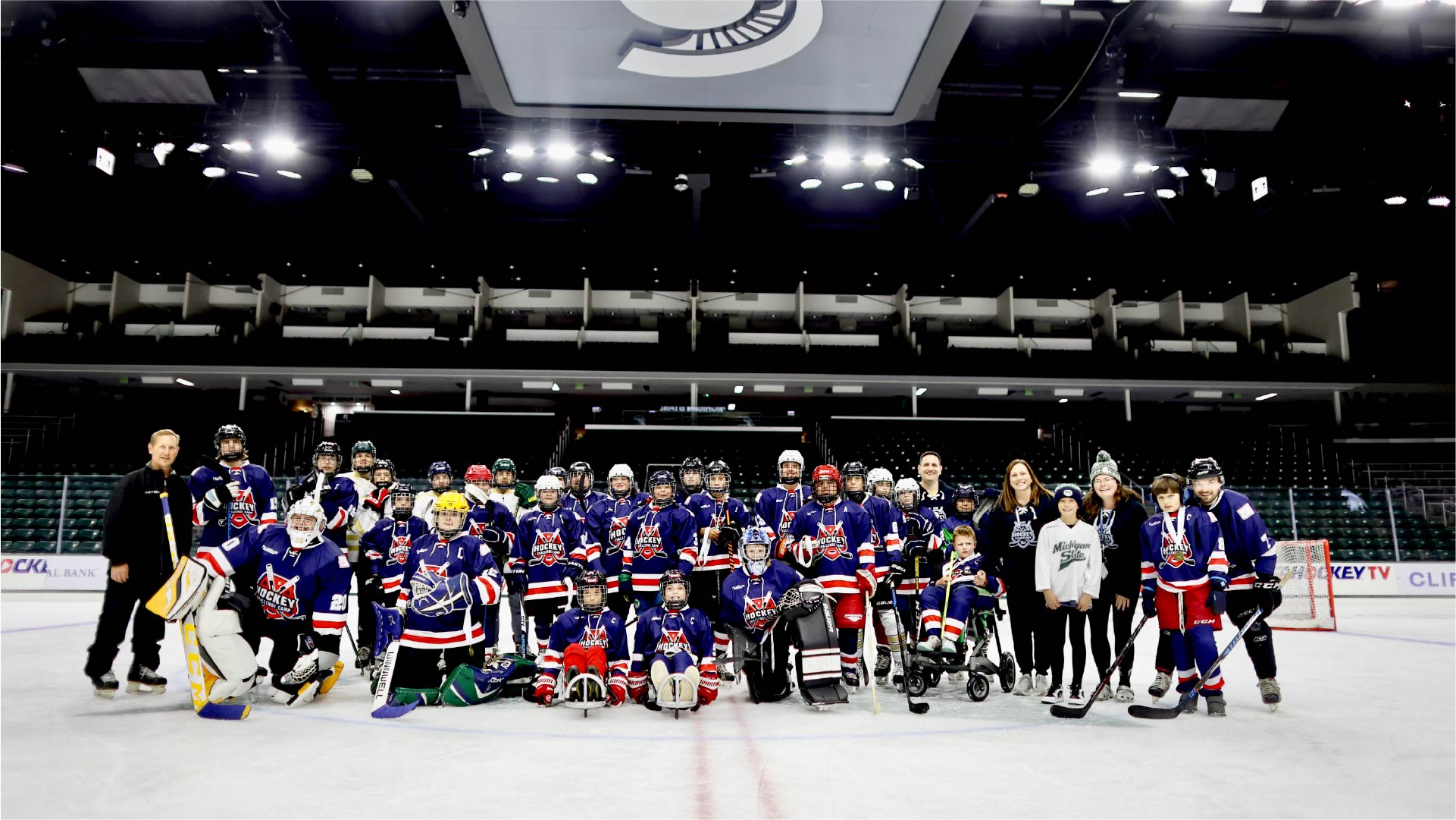 A large group of hockey players in blue jerseys with red and white accents poses together on an indoor ice rink. Some players are standing while others kneel in front, and several wear helmets and pads. A few individuals in casual clothing stand alongside the team. The background shows stadium seating and bright overhead lights, with a large Spartan logo displayed on the ceiling.