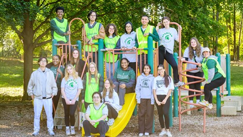 A large group of people wearing green safety vests and Spartan-themed shirts poses on and around a playground structure with slides and climbing bars. The background features tall trees and a grassy area.