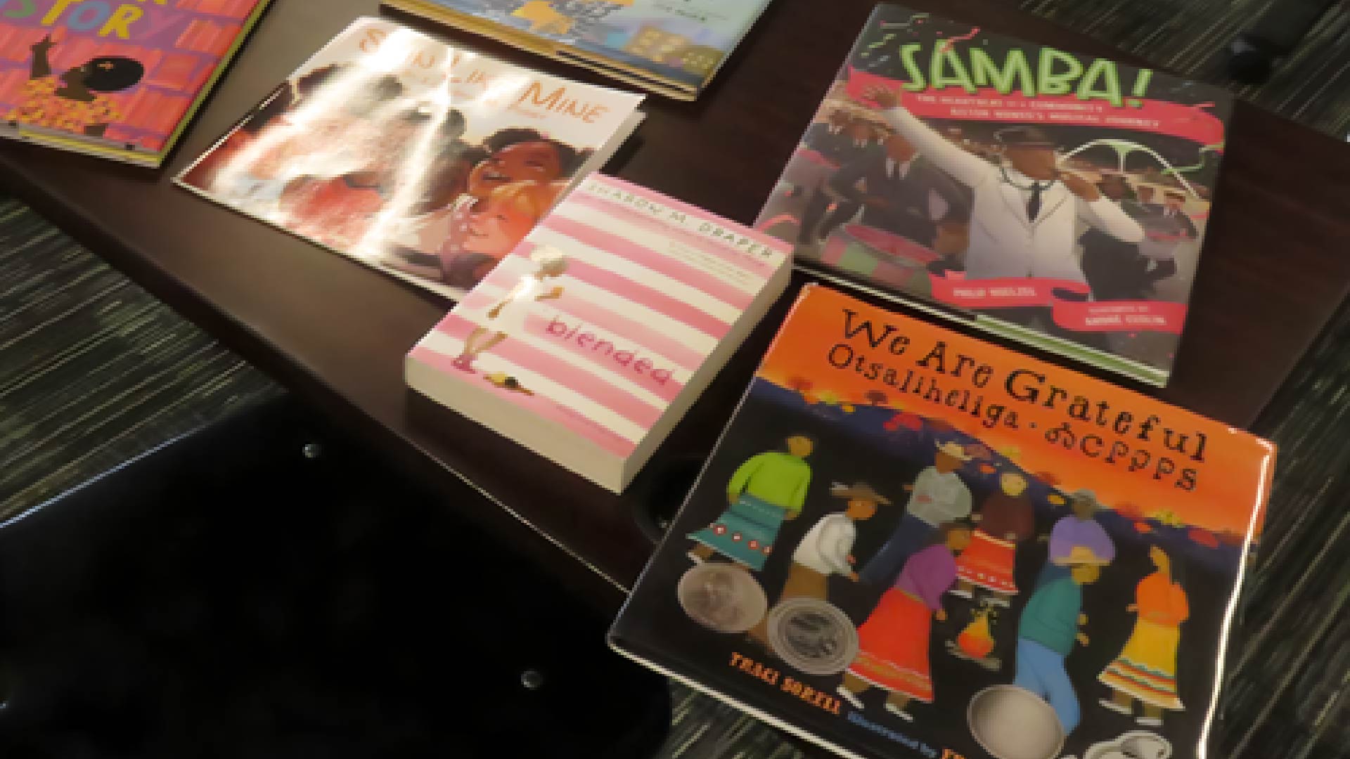A tabletop display of several colorful children’s books, including titles such as “We Are Grateful,” “Blended,” “Samba!” and others. The books are arranged neatly on a dark wooden surface.