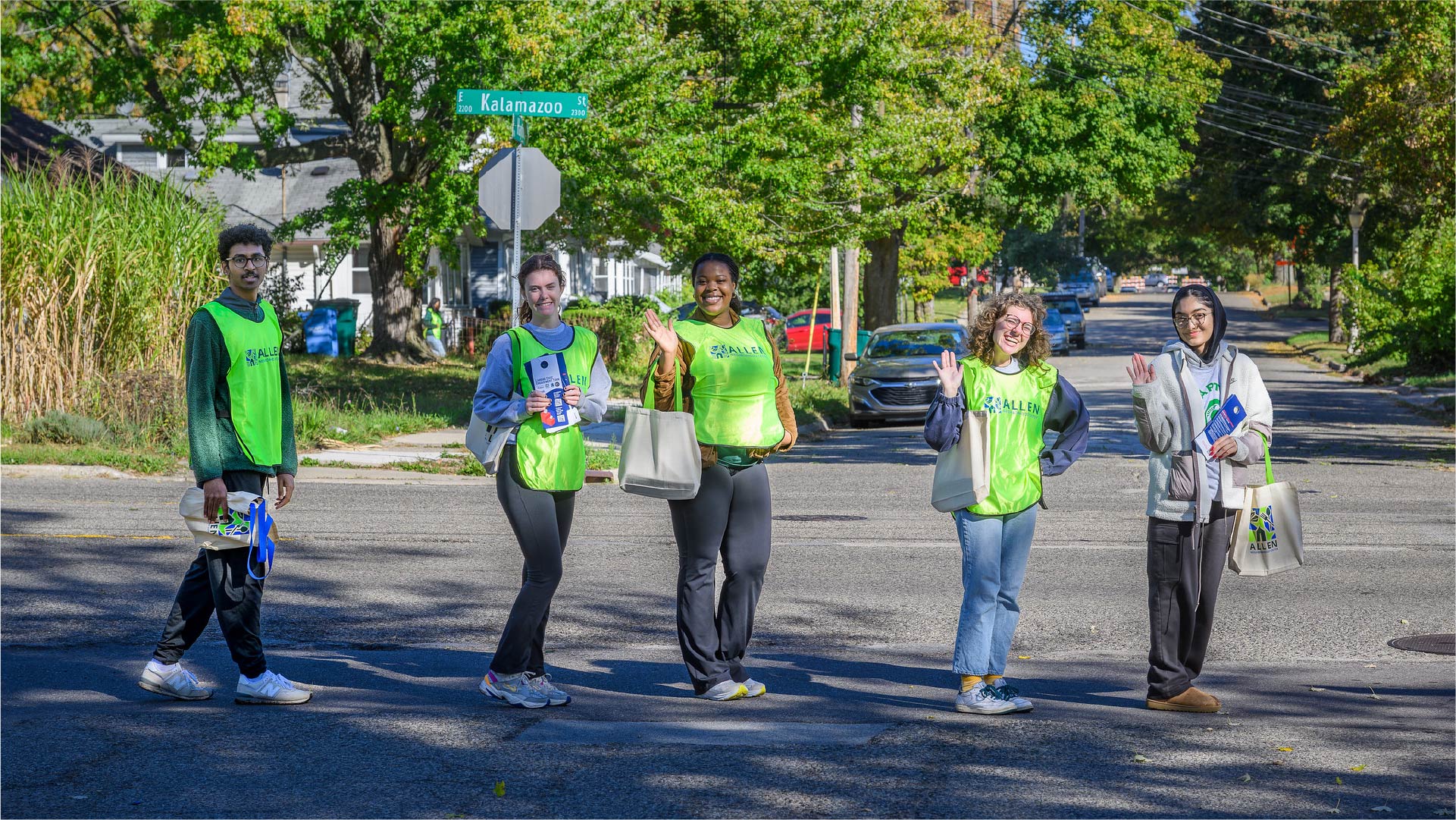 Five individuals wearing bright green vests stand in the middle of a residential street near a street sign labeled “Kalamazoo.” Each person carries tote bags and informational materials. Trees with green foliage and parked cars line the background.
