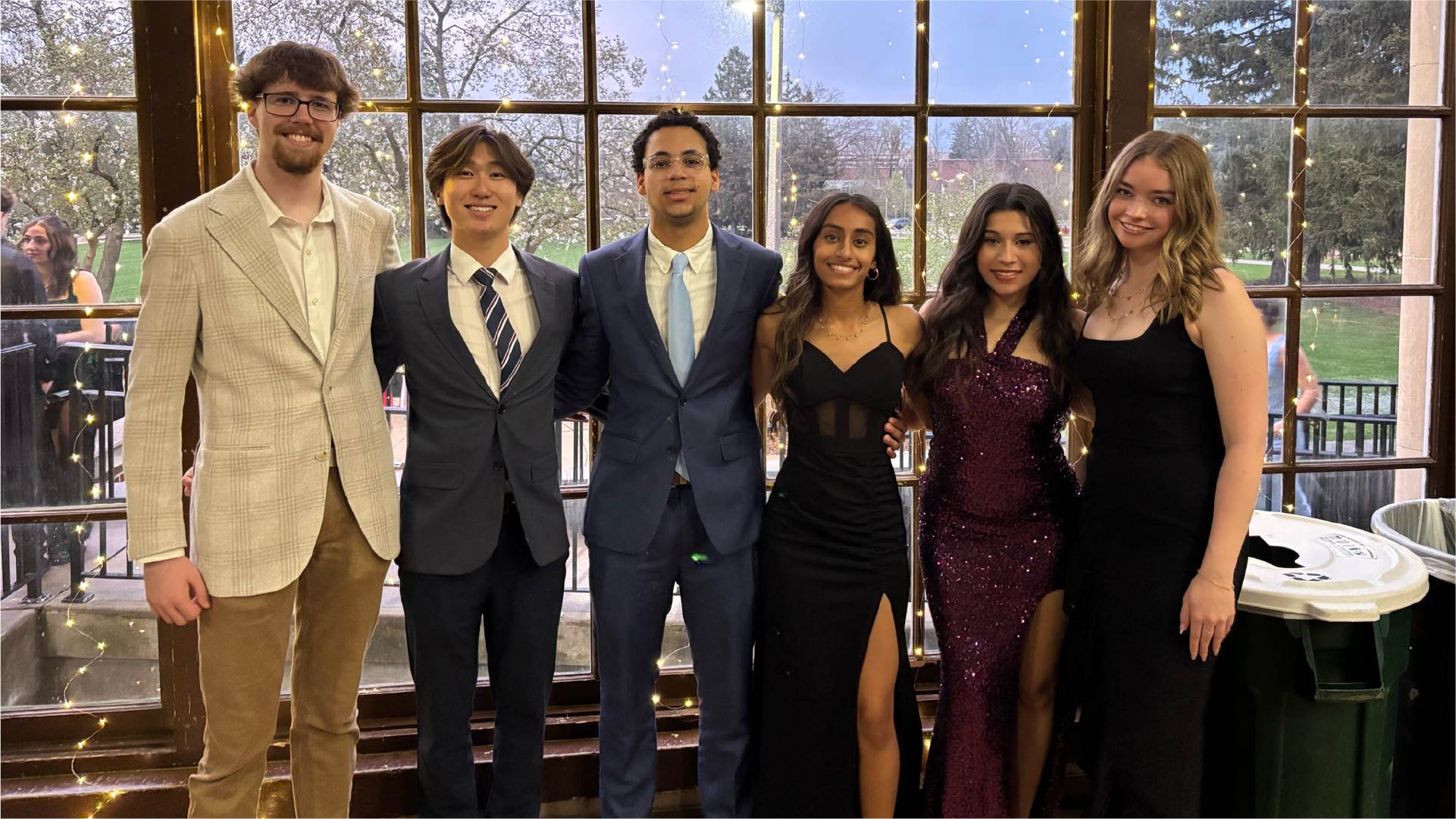 Six individuals stand side by side in front of large windows decorated with string lights. The group is dressed in formal attire, including suits and evening gowns. Outside the windows, trees and a lawn are visible at dusk.