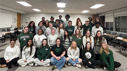 A large group of people poses together in a classroom setting. Most are wearing Michigan State University apparel in green, white, and gray. The room has long tables, chairs, and a whiteboard in the background with writing on it.