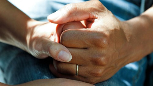 Close-up of two hands clasped together in a comforting gesture. One hand is wearing a simple gold ring on the ring finger. The background is softly blurred, showing blue fabric, likely clothing. Natural light highlights the texture and details of the skin.