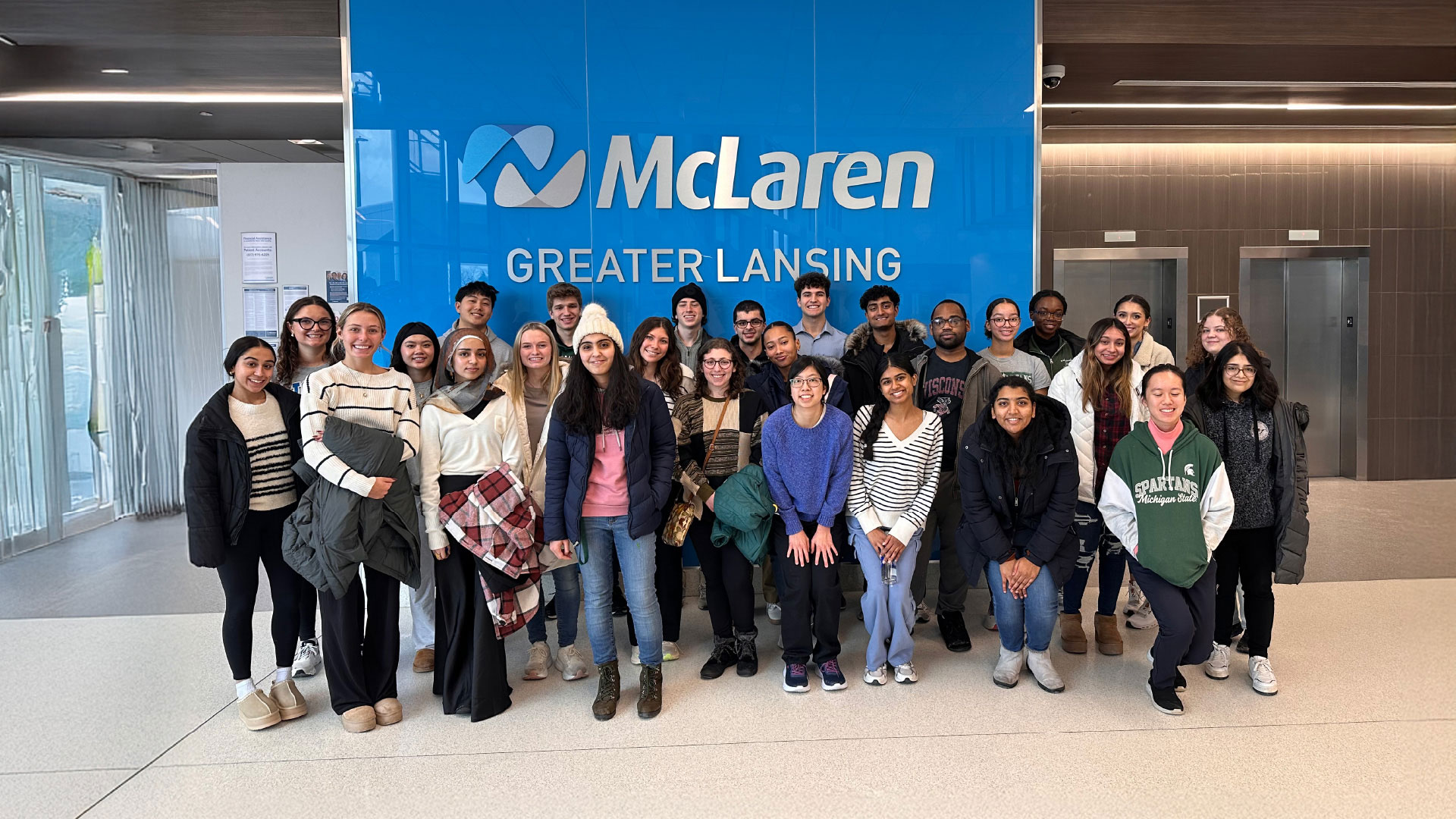 A group of people stands in front of a bright blue wall with the McLaren Greater Lansing logo. The setting appears to be the lobby of a modern building with elevators and glass doors.