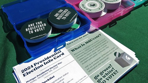 Close-up of a table with election-related materials, including circular stickers that read “Are you registered to vote?” and informational cards about the 2024 Presidential Election and MSUVote. The items are arranged in colorful plastic containers.