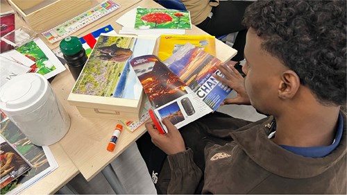 An individual sits at a table working on a collage using magazine pages. The table has glue sticks, a water bottle, and colorful images of landscapes and flowers arranged on wooden boards.