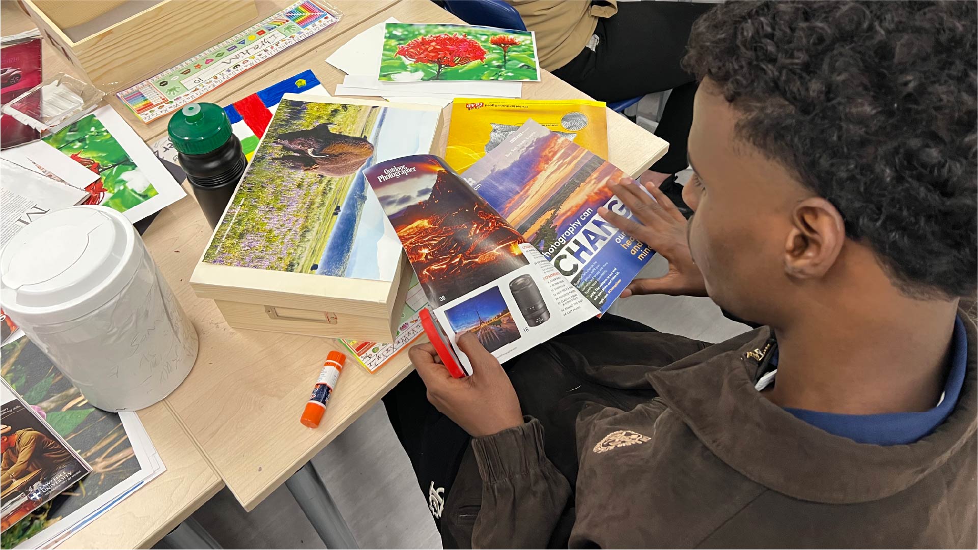 An individual sits at a table working on a collage using magazine pages. The table has glue sticks, a water bottle, and colorful images of landscapes and flowers arranged on wooden boards.