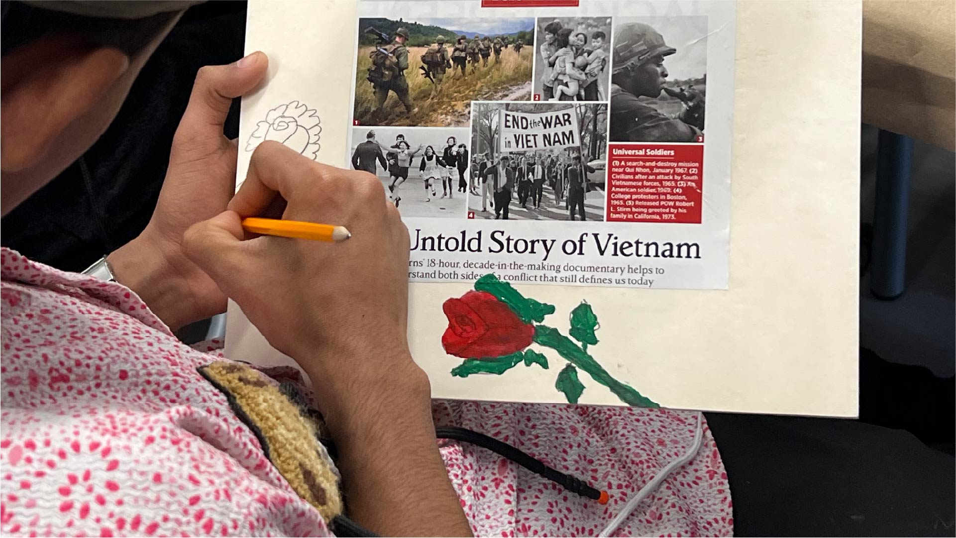 A person works on a collage featuring historical photos and text about the Vietnam War. The page includes the headline “Untold Story of Vietnam” and a red rose illustration being drawn with a pencil.