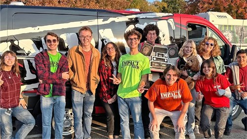 A group of people stands in front of a black and red van decorated with a large graphic of a boombox and the words “Impact 89FM.” Several individuals wear bright green or red shirts with the station’s logo, and autumn trees are visible in the background.