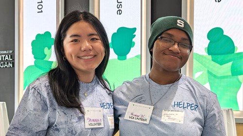 Two volunteers wearing light blue shirts with “HELPER” printed on them stand side by side indoors. They have name tags and are in front of a display with green graphics.