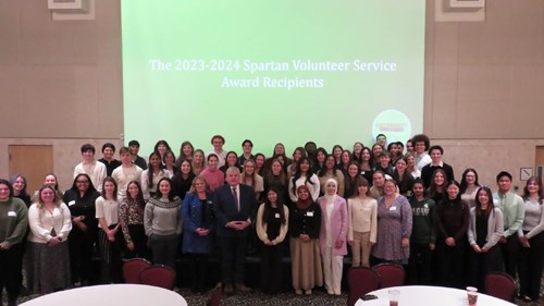 A large group of people posing in front of a projection screen that reads “The 2023-2024 Spartan Volunteer Service Award Recipients.” Round tables with drinks and papers are visible in the foreground.