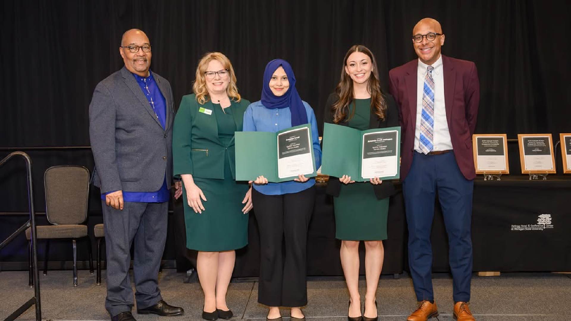 Five individuals standing on a stage holding certificates. Plaques are displayed on a table in the background. The setting appears to be a formal awards event with a black backdrop.