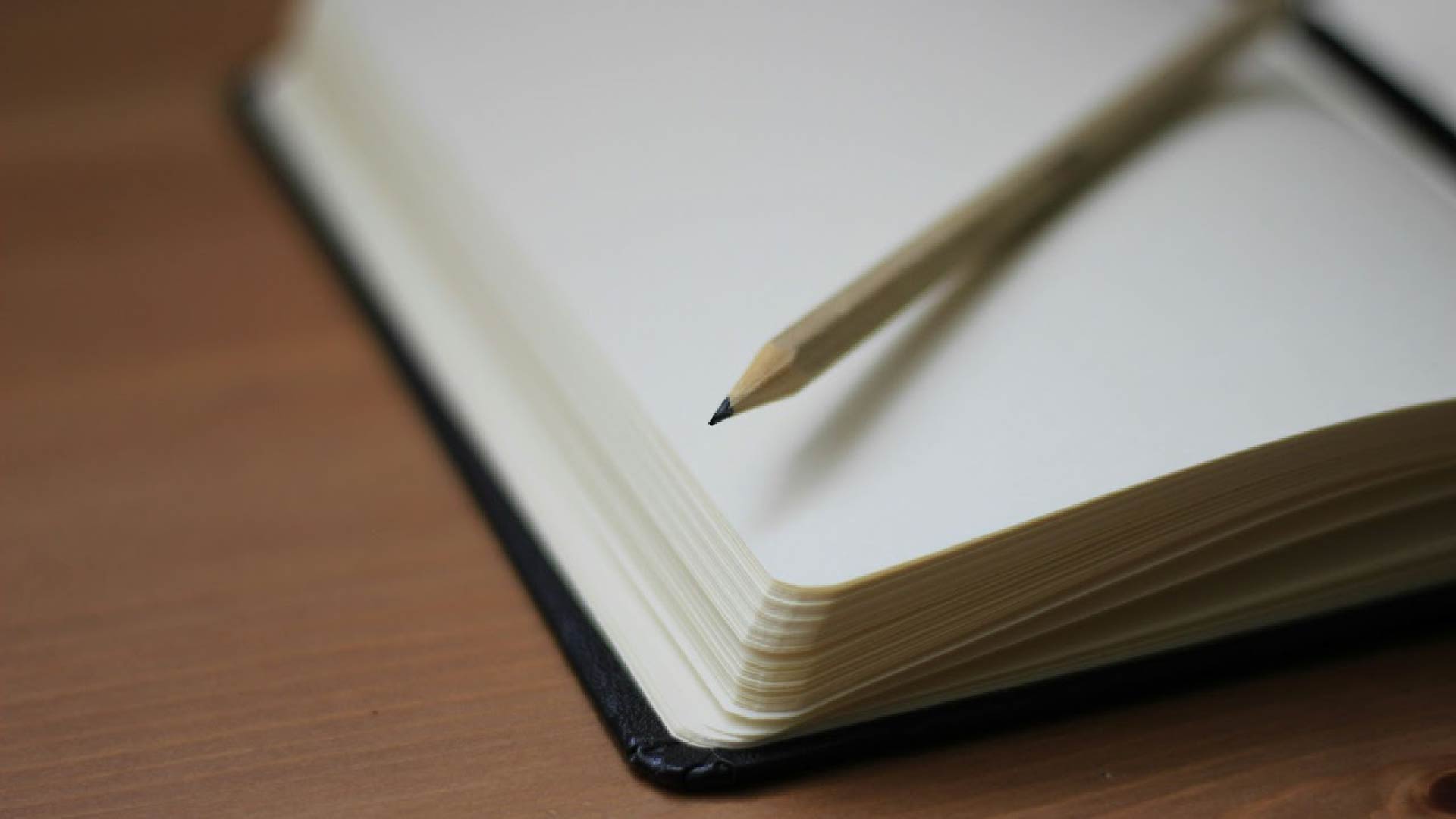 A close-up of an open blank notebook with a wooden pencil resting diagonally across the pages. The notebook is placed on a wooden surface.