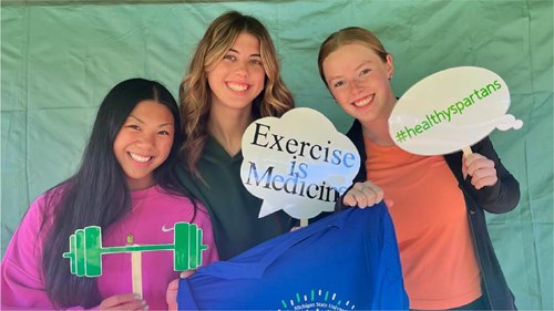 Three individuals stand in front of a green backdrop holding props. One prop is shaped like a barbell, another is a speech bubble reading “Exercise is Medicine,” and the third says “#healthyspartans.” They are also holding a blue T-shirt with a logo.