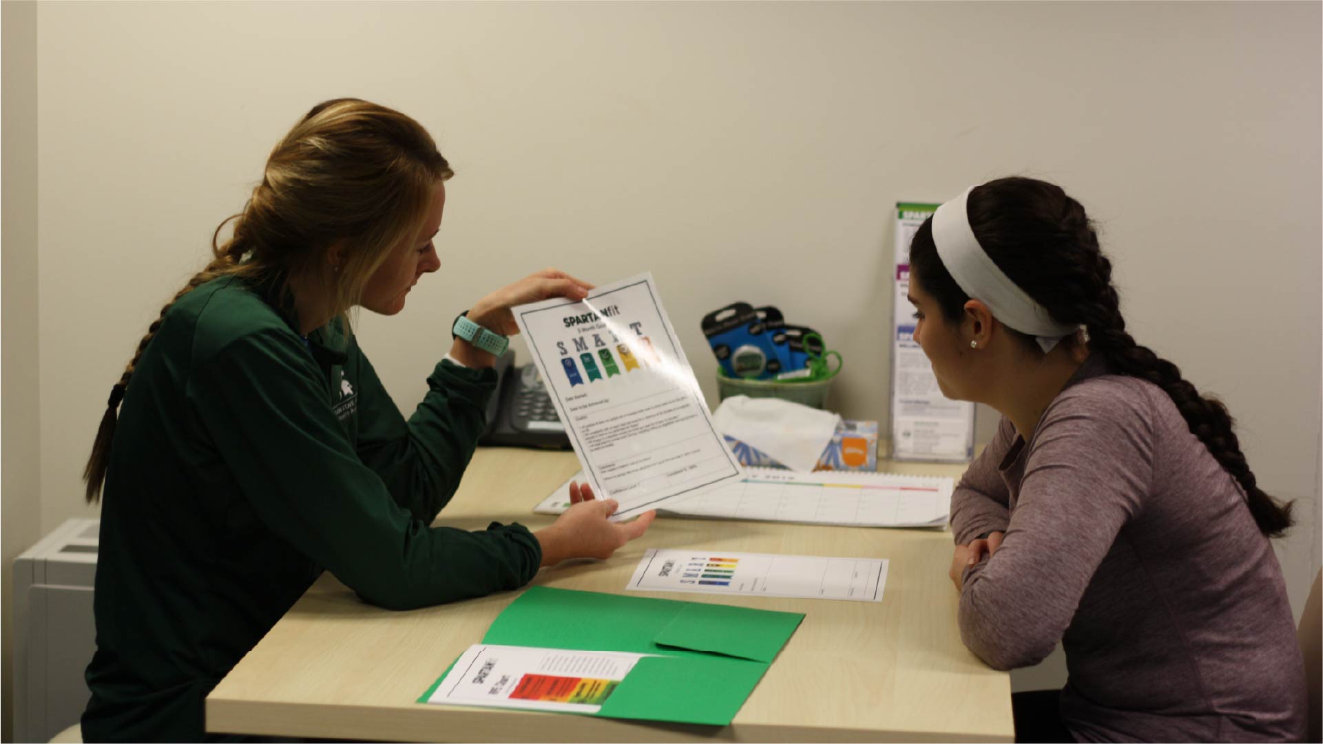 Two individuals sit at a table in a small office setting. One person is holding and pointing to a printed sheet titled “SPARTAN MOVE” with colorful sections, while the other listens attentively. The table has a green folder, papers, and a ruler, and exercise equipment is visible in the background.