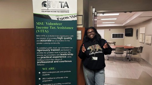 A person stands next to a tall banner for MSU Volunteer Income Tax Assistance (VITA). The banner includes text about the program’s mission and services. The setting is an indoor room with tables and chairs.