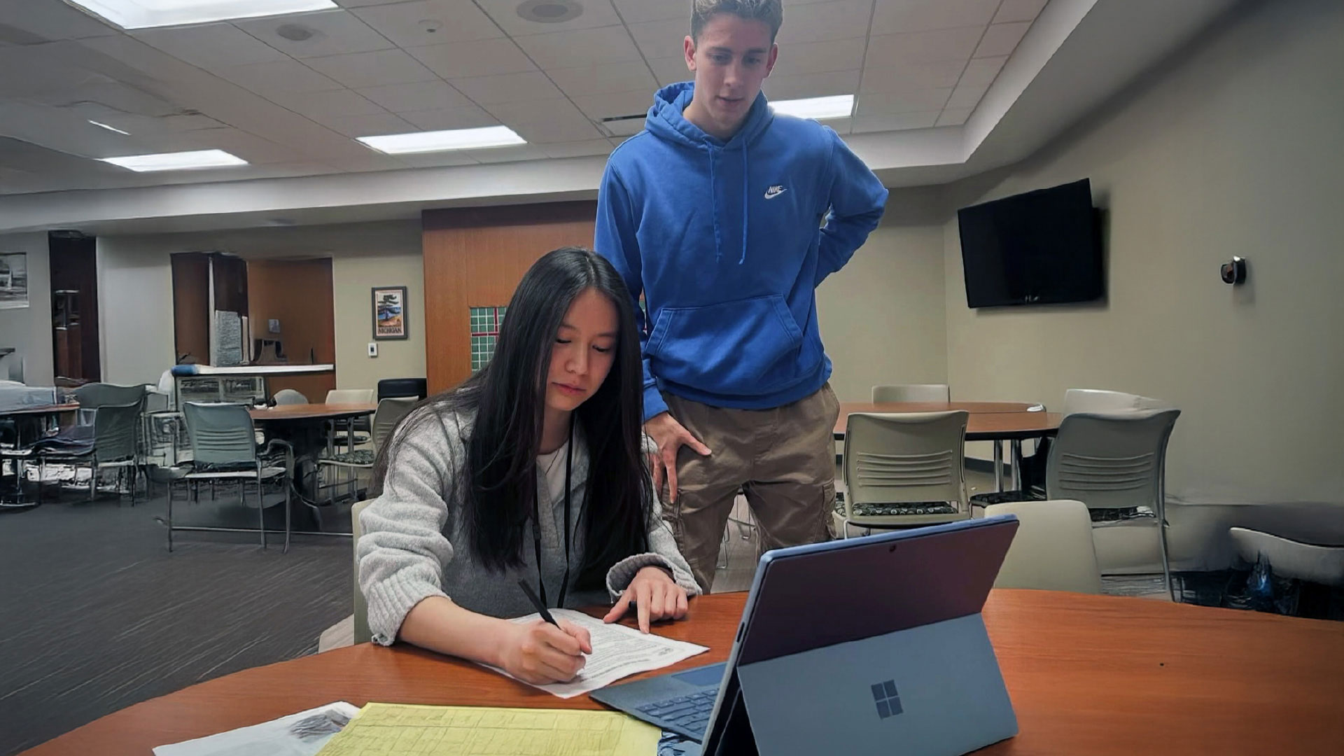 Two individuals are in a room with tables and chairs. One person is seated, writing on a form next to a laptop, while the other stands nearby observing.