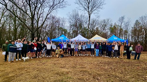A group photo of Interfraternity Council students and community volunteers at a River Trail beautification site with Michigan Waterways Stewards tents in the background during a cleanup event.