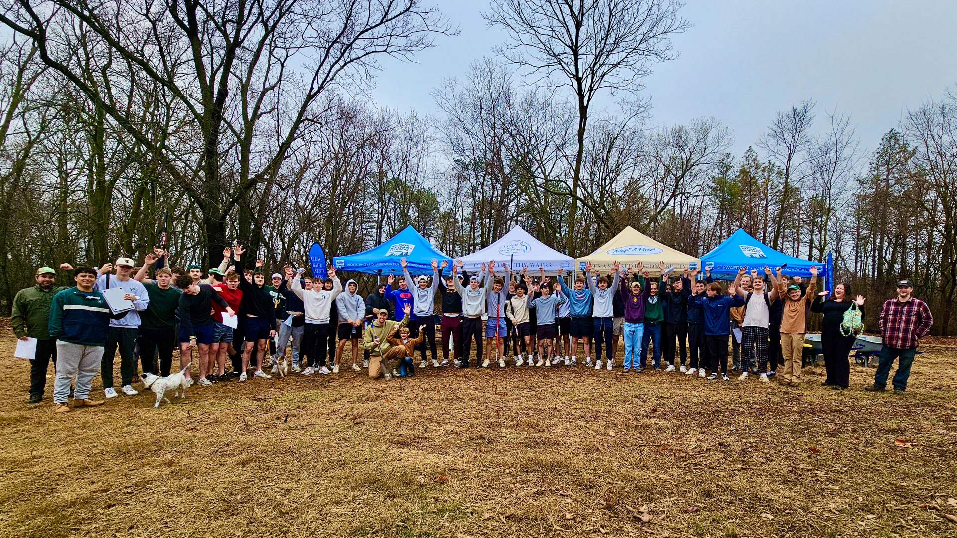 A group photo of Interfraternity Council students and community volunteers at a River Trail beautification site with Michigan Waterways Stewards tents in the background during a cleanup event.
